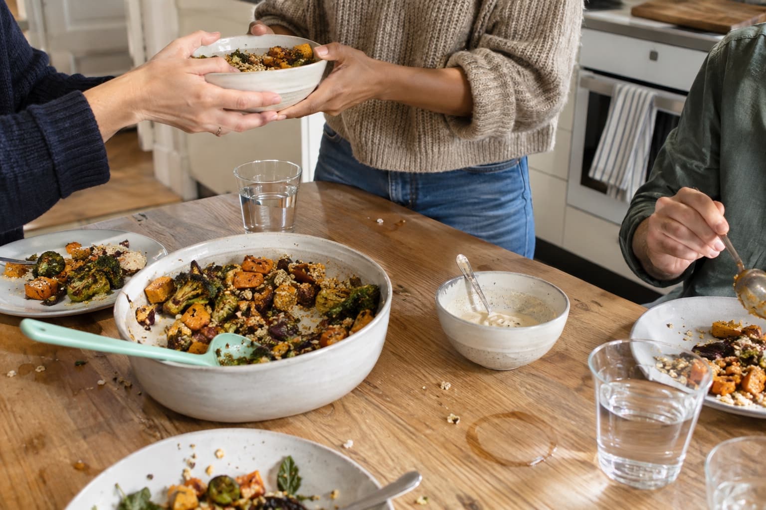 Three friends passing colourful plant-based food around a well-used kitchen table