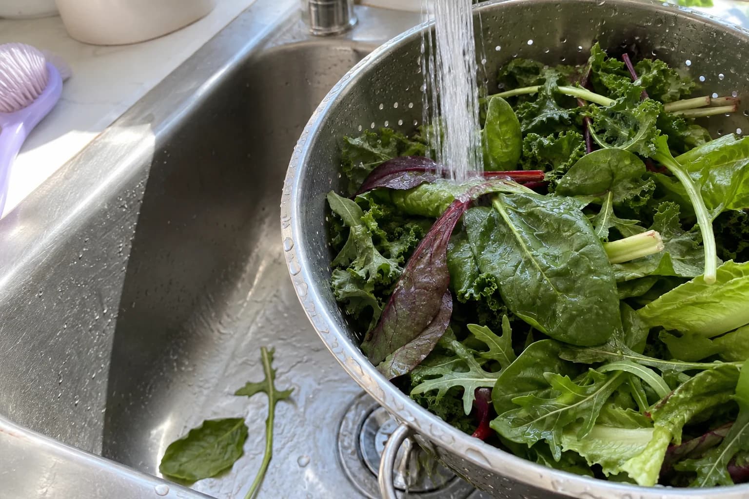 Mixed leafy greens being rinsed in a metal colander in an ordinary kitchen sink