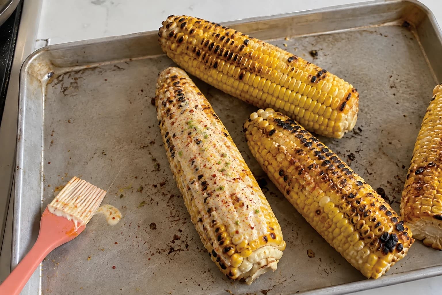 Four irregularly charred corn cobs on a used tray, with one partly coated in a creamy chili-lime sauce