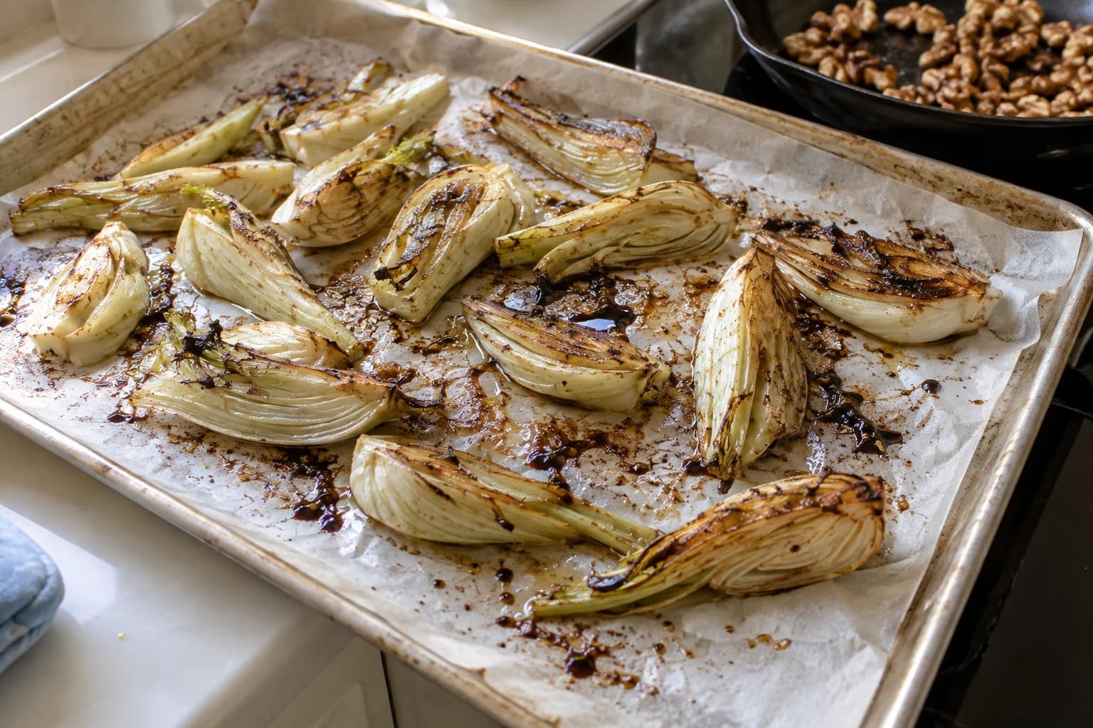 Caramelized fennel wedges roasting on a used sheet pan beside a small skillet of walnuts