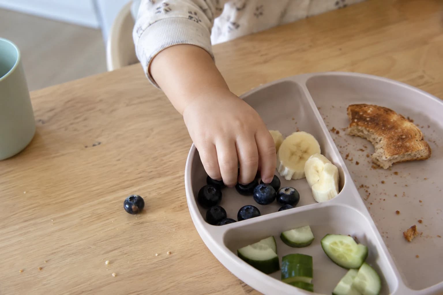 A toddler hand choosing blueberries from an ordinary snack plate at the kitchen table